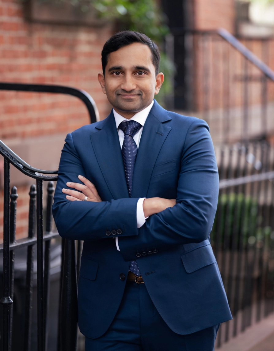 Karl Coutinho MD in navy blue suit standing with arms crossed outdoors near brick building and iron railing - in Clifton, NJ
