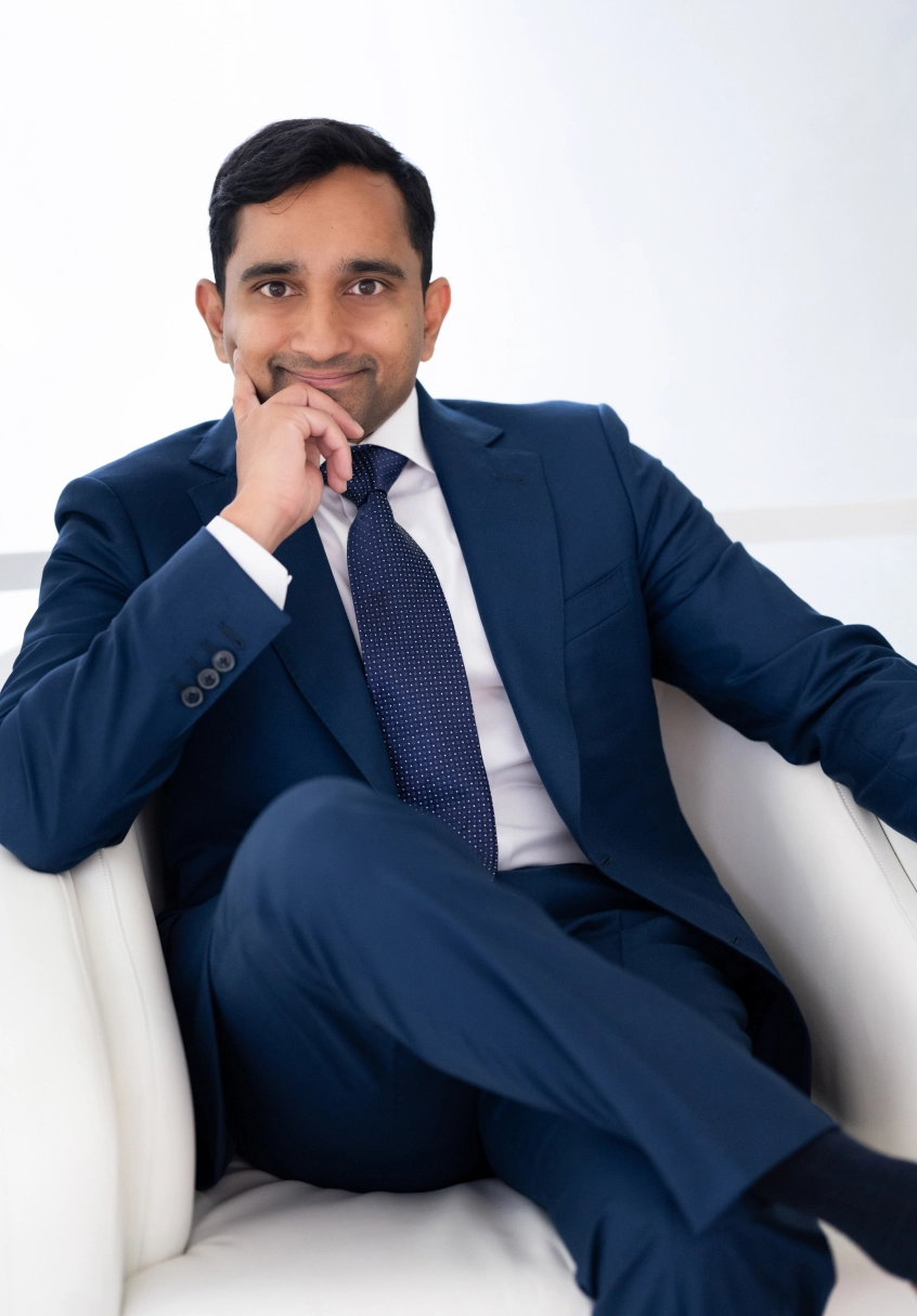Karl Coutinho MD in navy suit sitting thoughtfully in white chair against white background - Doctor in Clifton, NJ