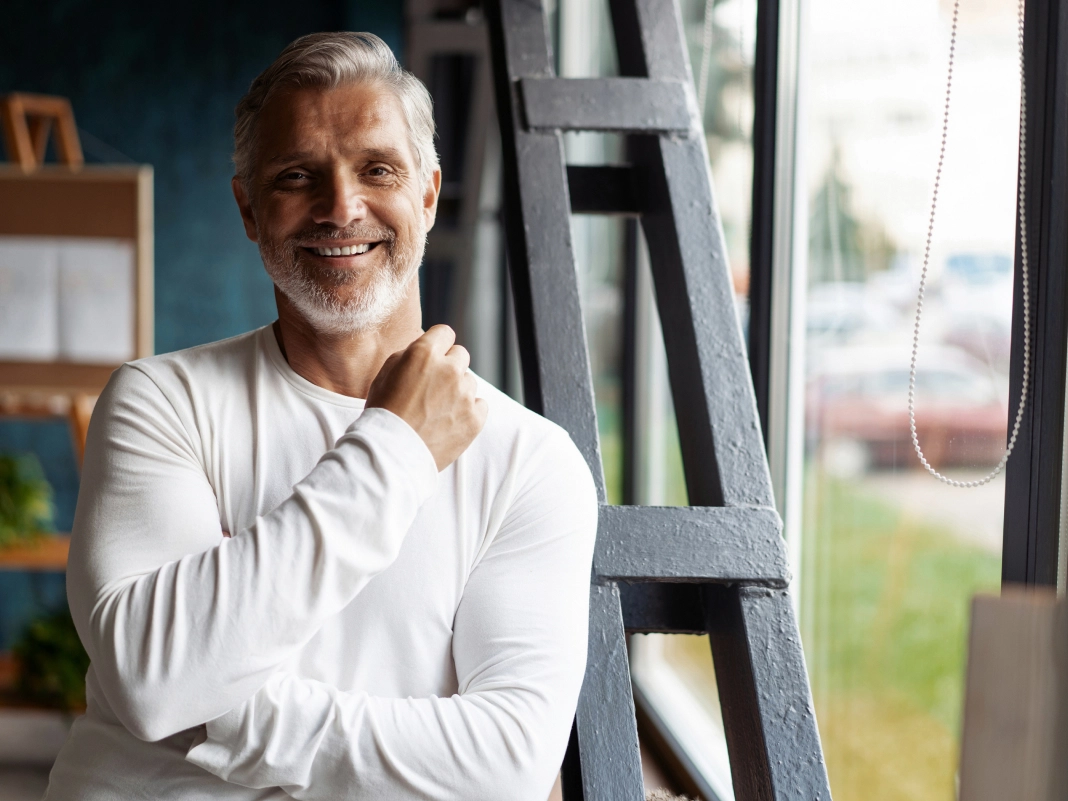 Smiling mature man with gray beard in white sweater standing confidently next to black ladder in bright modern interior - in Clifton, NJ