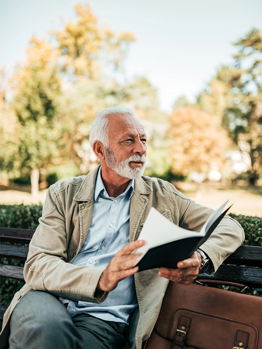 Senior man with white beard reading book on park bench in autumn setting - Prostate enlargment in Clifton, NJ