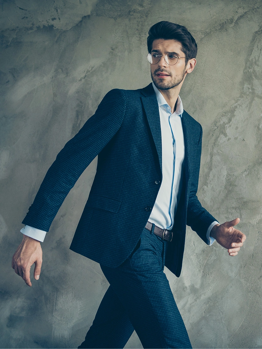 Young man in navy blue suit and white shirt walking against textured grey background in fashion portrait - scrotoplasty in Clifton, NJ