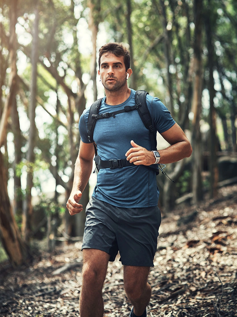 Man trail running through forest wearing blue shirt and backpack on wooded path - vasectomy in Clifton, NJ