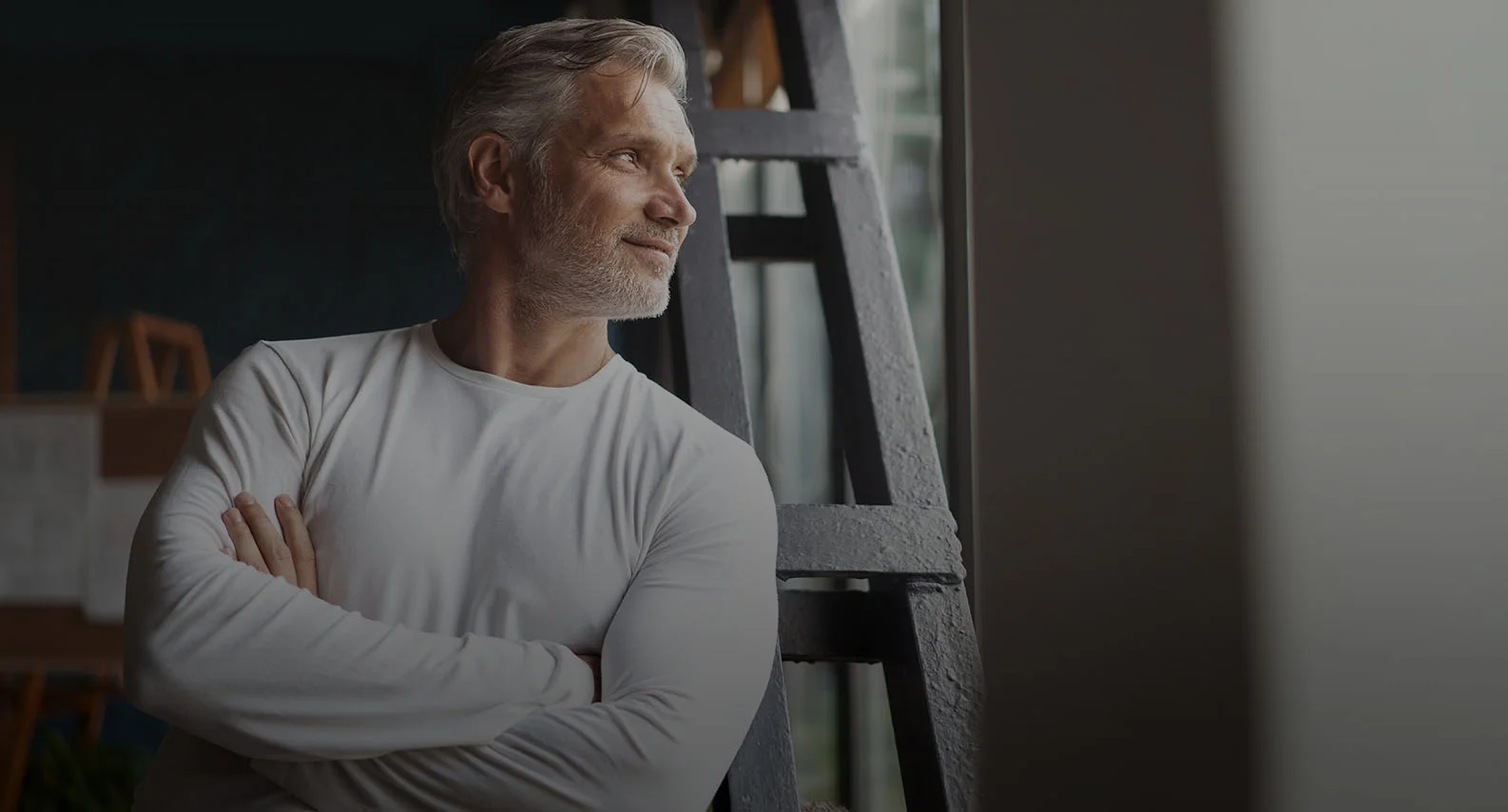 Confident man in white long-sleeve shirt with arms crossed looking pensively out window in modern interior - in Clifton, NJ