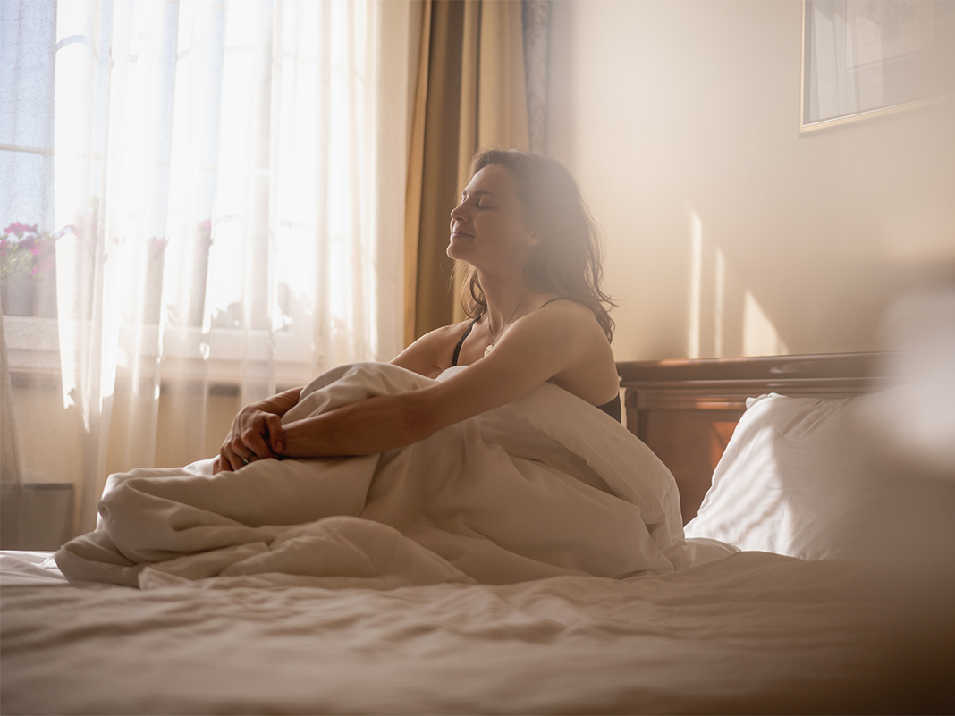 Woman sitting on hotel bed in bright sunlight looking out window during medical travel stay - hotels in Clifton, NJ
